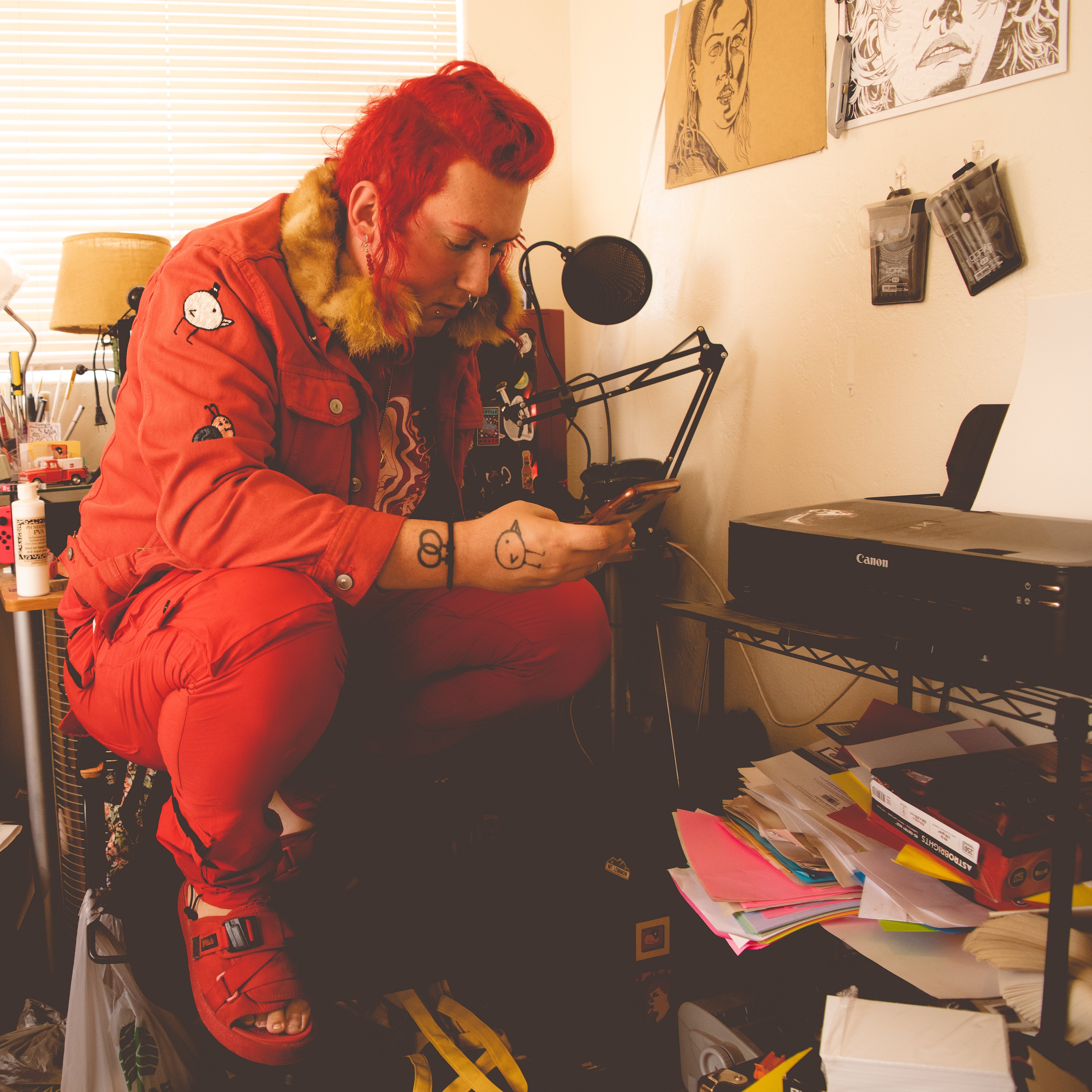 a picture of ruby may valentine, she is wearing entirely red clothes that look very cool, her hair is also entirely red, shes crouched on a chair looking at her phone in a cluttered room